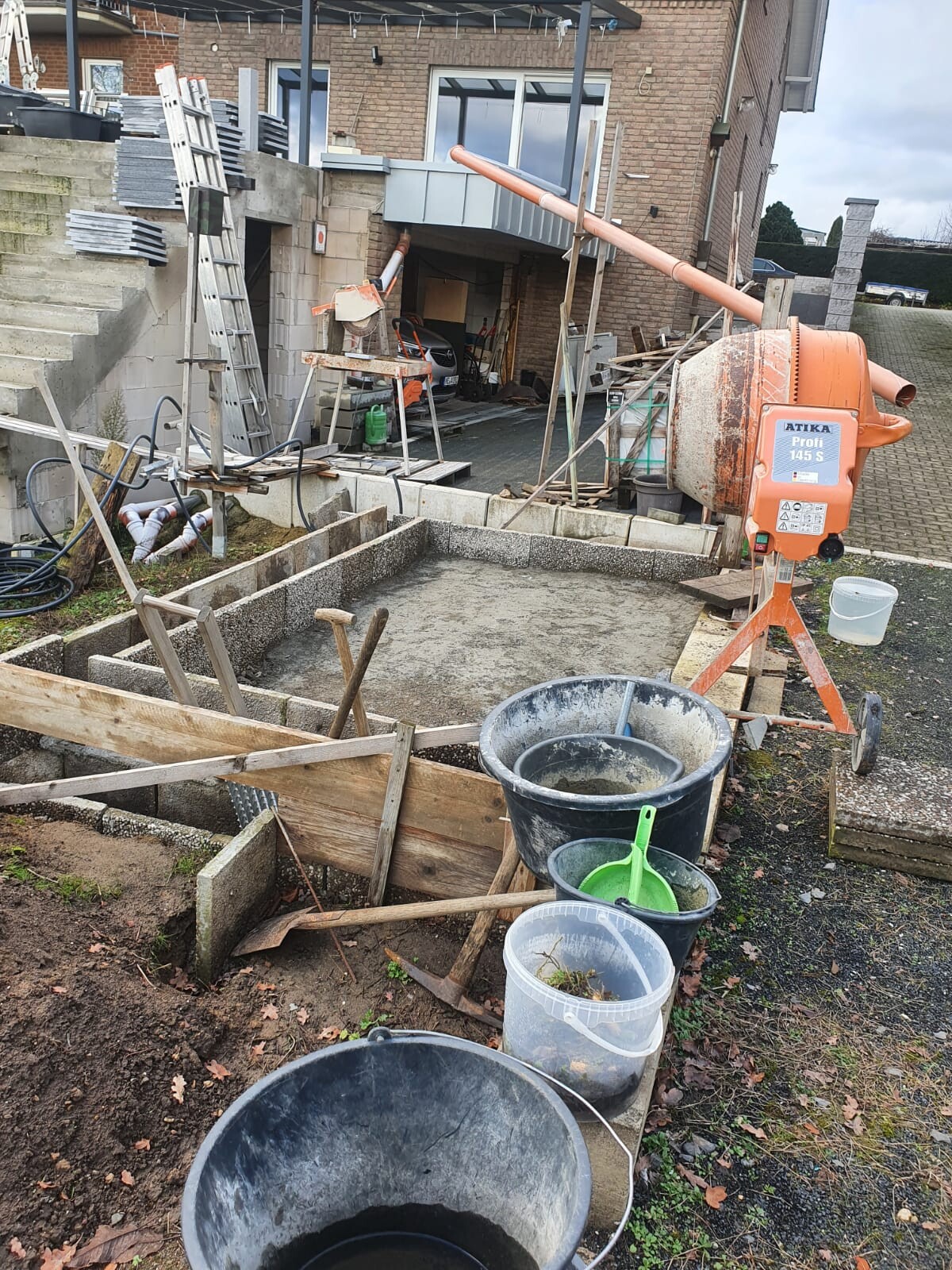 Baustelle vor einem Wohnhaus: Betonmischer, Bretter, Eimer, Schubkellen und Bauarbeiten.