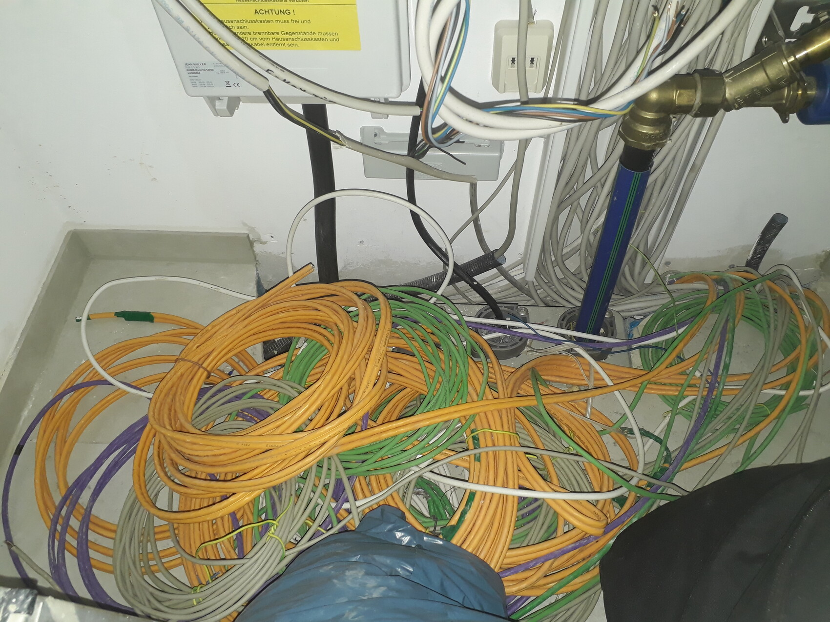 Verknotete bunte Kabel (orange, grün, lila) auf dem Boden neben Rohren und Geräten.