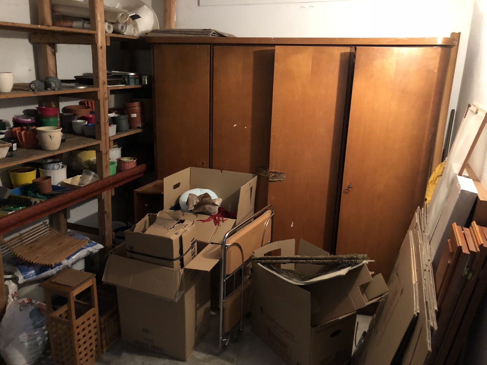 Abstellraum mit großem Holzmöbel-Schrank, Kartons, Regalen und bunten Blumentöpfen.