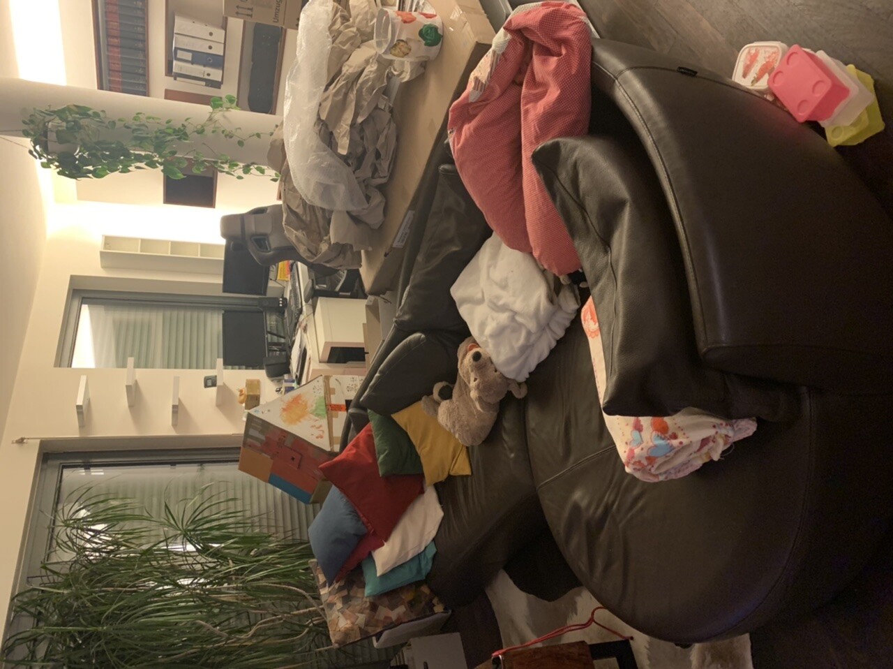 Wohnzimmer mit Ledercouch, Decken, Kissen und Spielzeug; Bürobereich im Hintergrund.