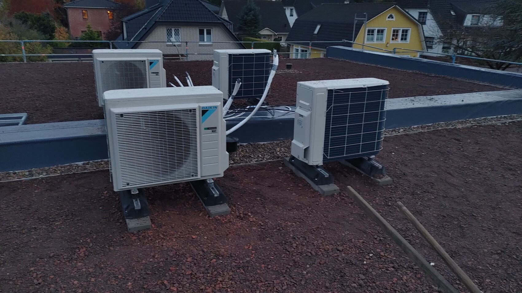 Vier Außengeräte einer Klimaanlage auf Flachdach mit Kabeln, Kiesboden, Häuser im Hintergrund.