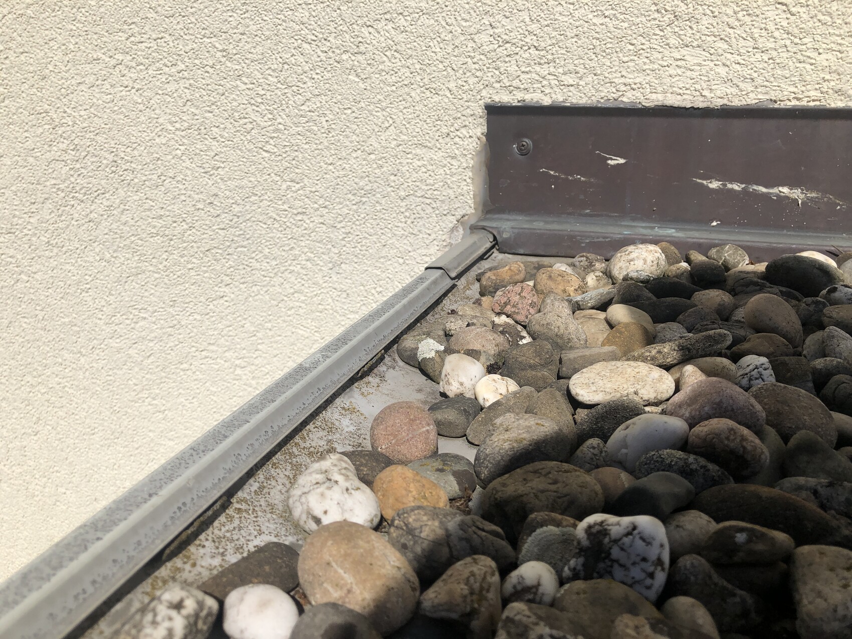 Beige verputzte Wand mit Kieselsteinen auf dem Dachrand und einem Metallprofil.