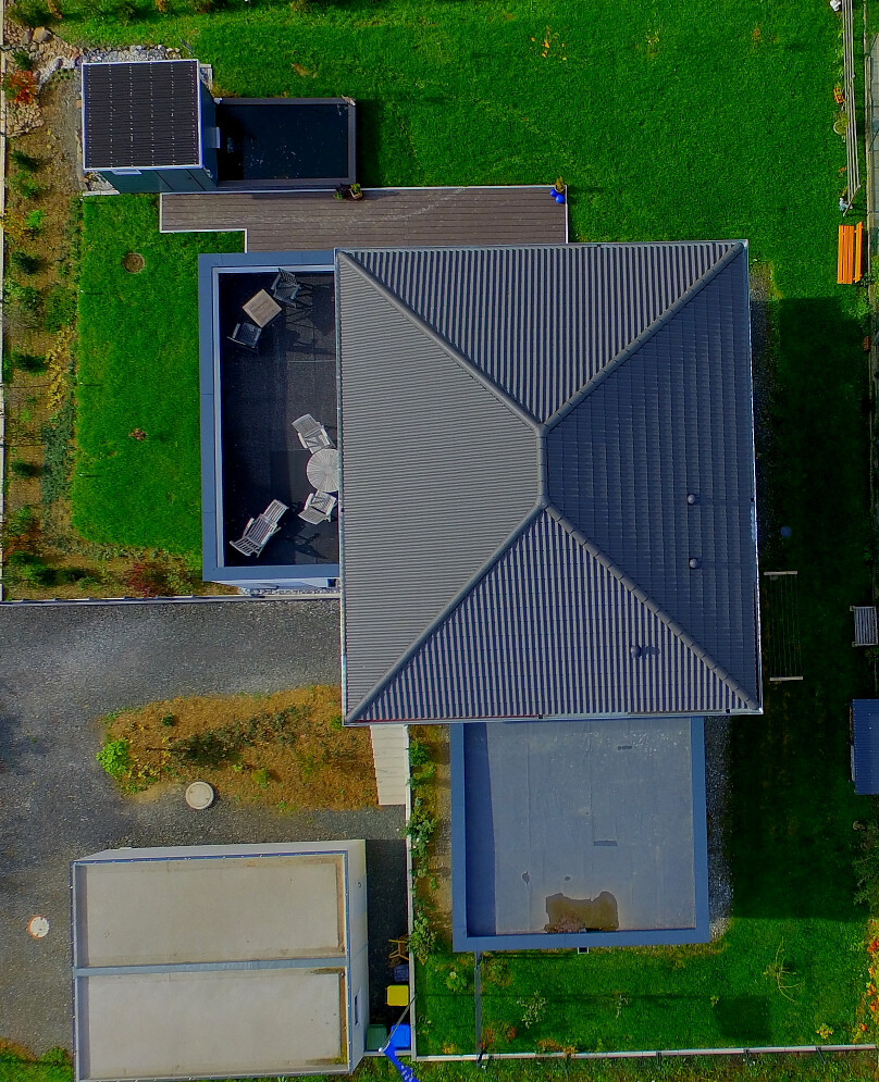 Vogelperspektive eines modernen Hauses mit großem, mehrteiligem Dach, Terrasse und grünem Garten.