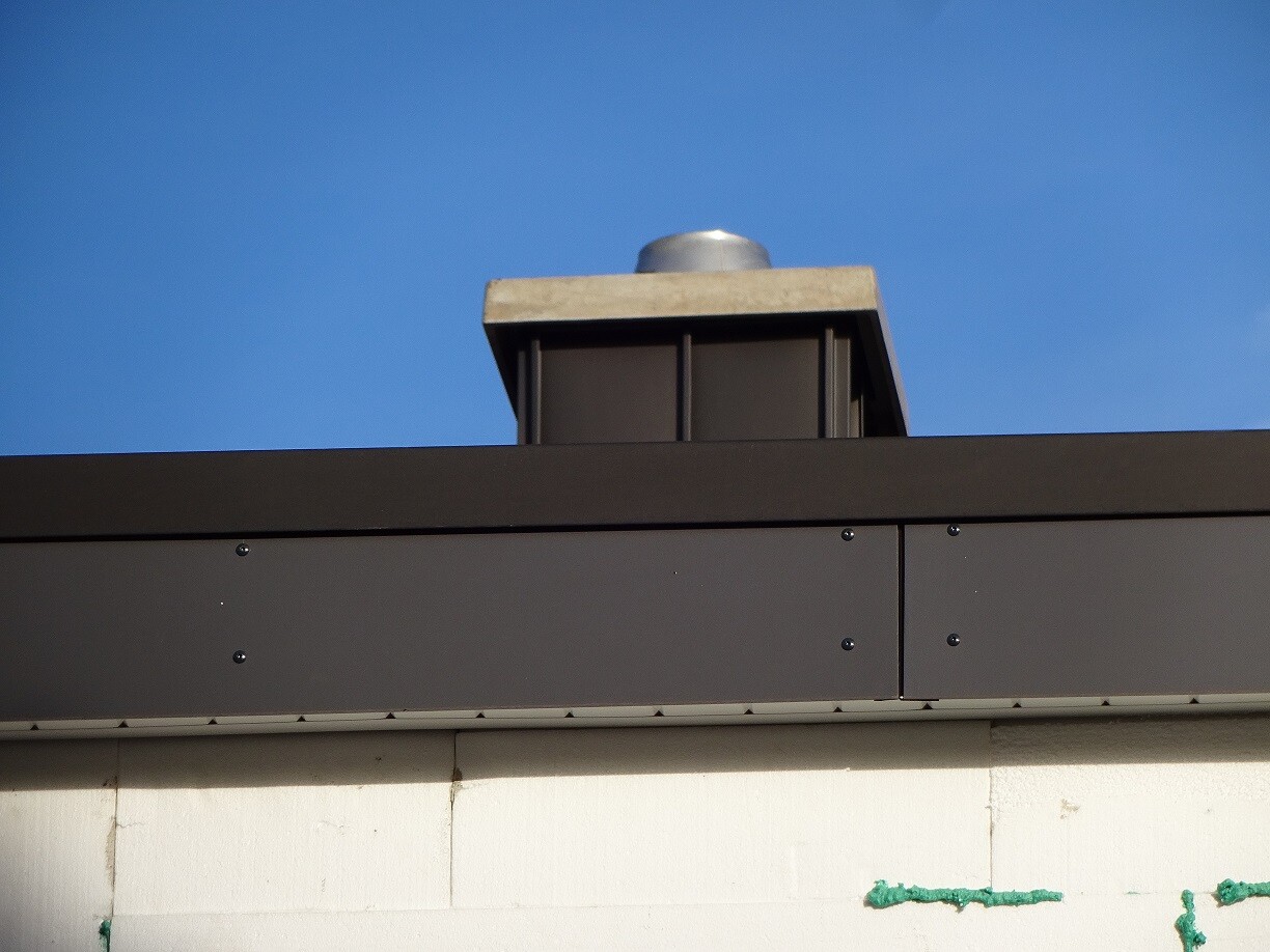 Draufsicht auf dunkle Metallverkleidung des Dachrandes mit einem runden Dachlüfter gegen blauen Himmel