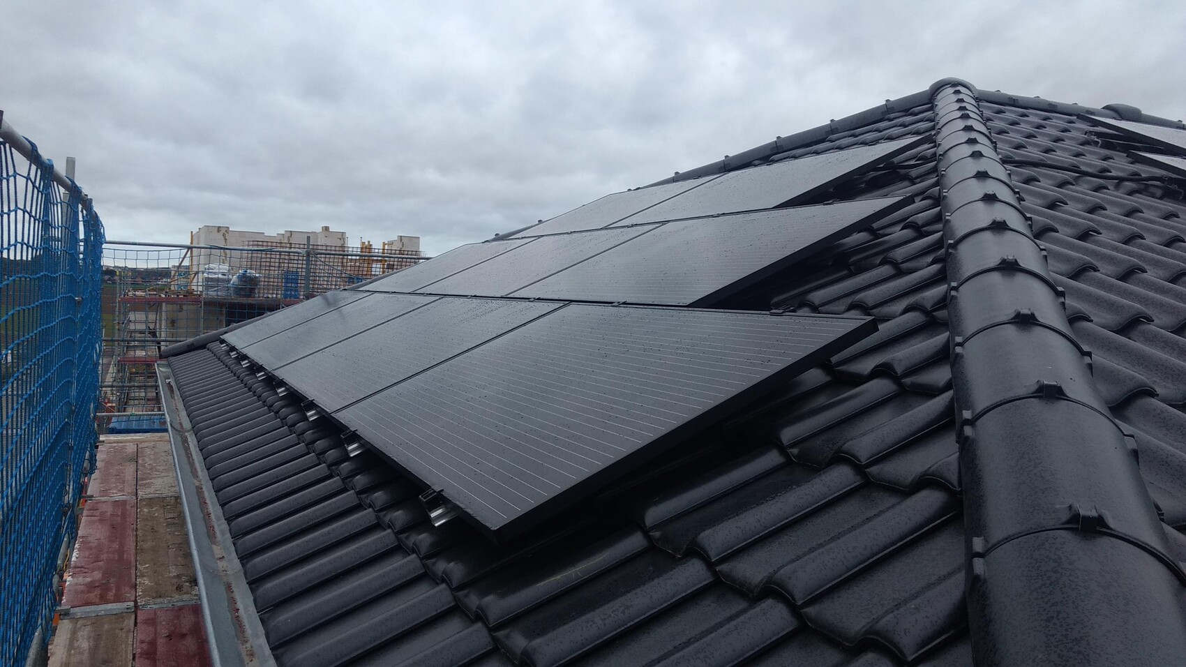 Dach mit Solarzellen auf dunklen Dachziegeln; Baugerüst und blaues Netz am Rand, wolkiger Himmel