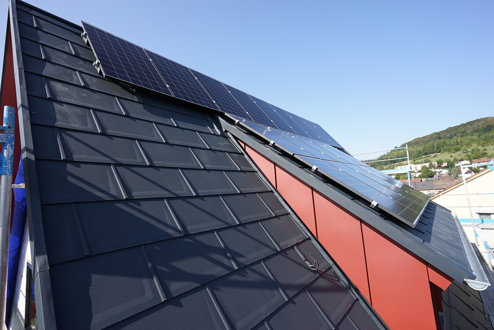 Dach mit schwarzen Schieferziegeln und Solarmodulen, rote Seitenwand, blauer Himmel.