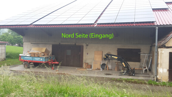Nordseite eines Scheunentrakts mit Solarpanel-Dach, Baugerät, Anhänger und Leiter am Eingang.