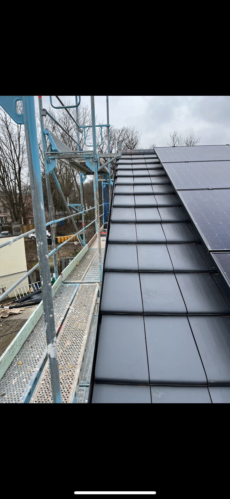 Dach mit schwarzen Ziegeln, rechts installierte Solarpaneele; links Gerüst.