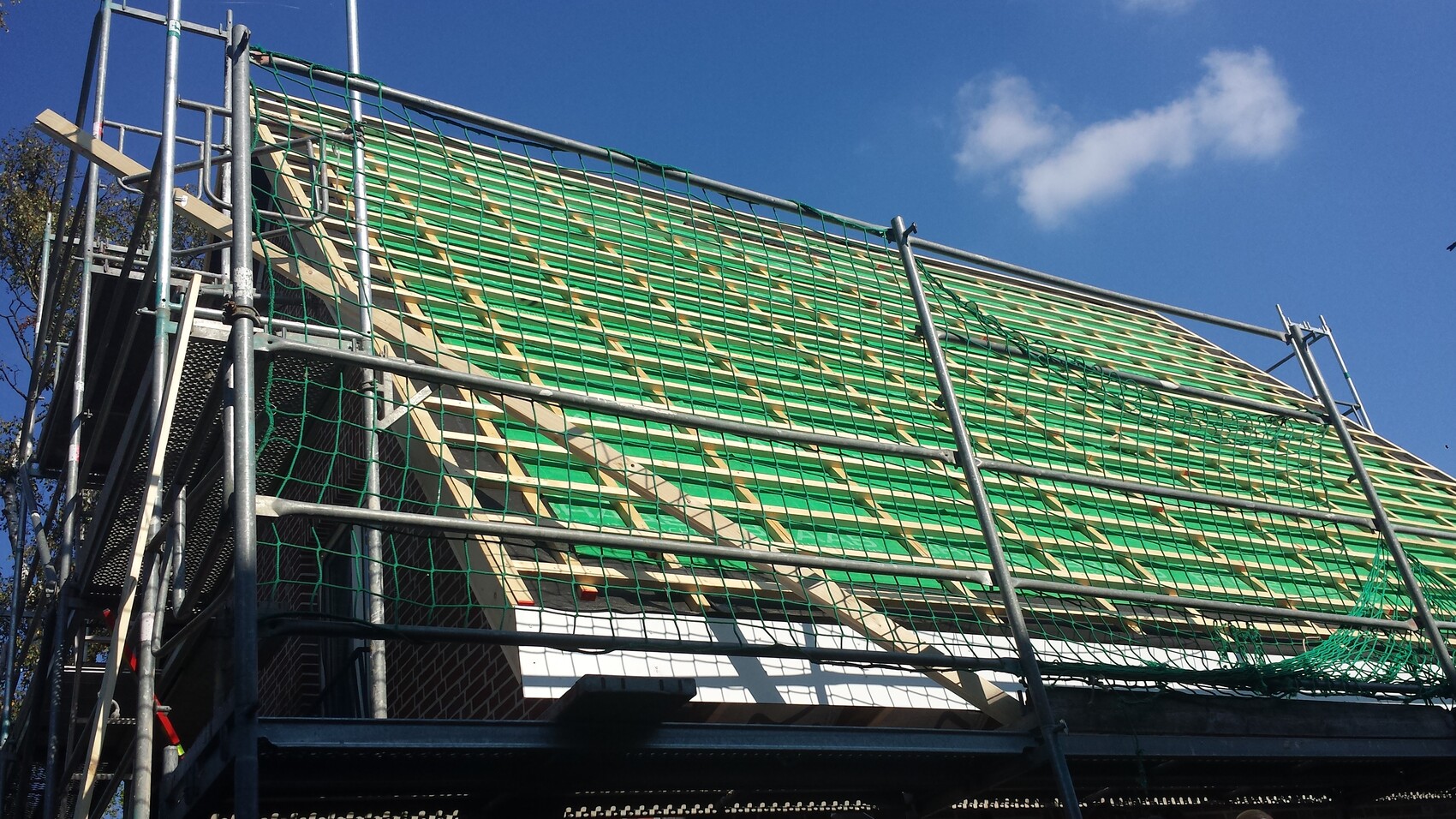 Dachkonstruktion mit grünem Unterdach, Holzlatten, Baugerüst mit Netz vor blauem Himmel