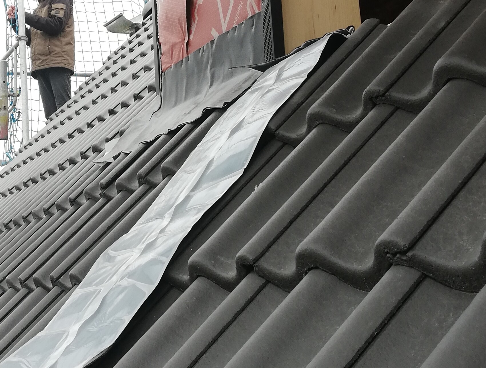 Dachdecker auf grauem Ziegeldach mit Plastikfolie, Gerüst im Hintergrund.