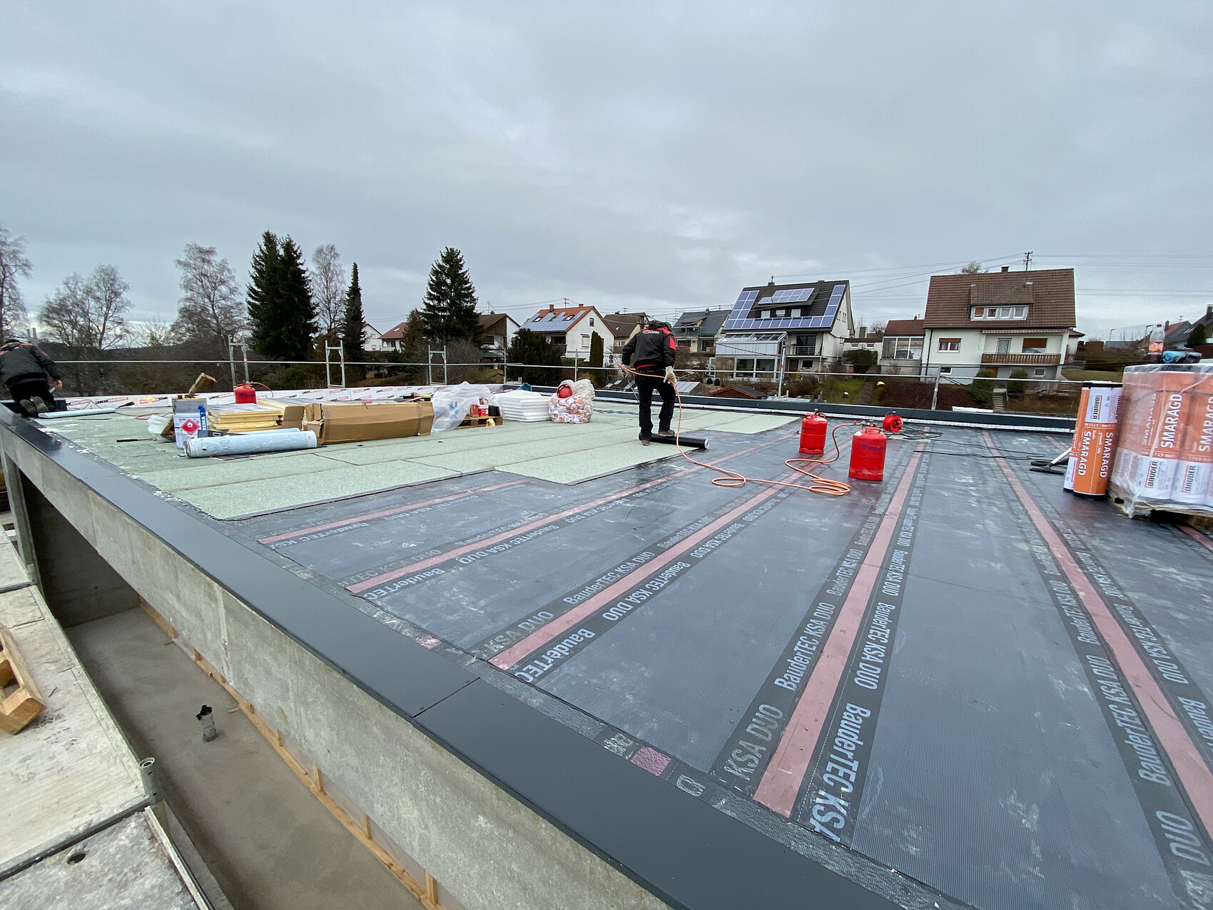 Auf einer Dachfläche arbeiten Bauarbeiter; Dämmung, Materialien und Gasflaschen liegen bereit.