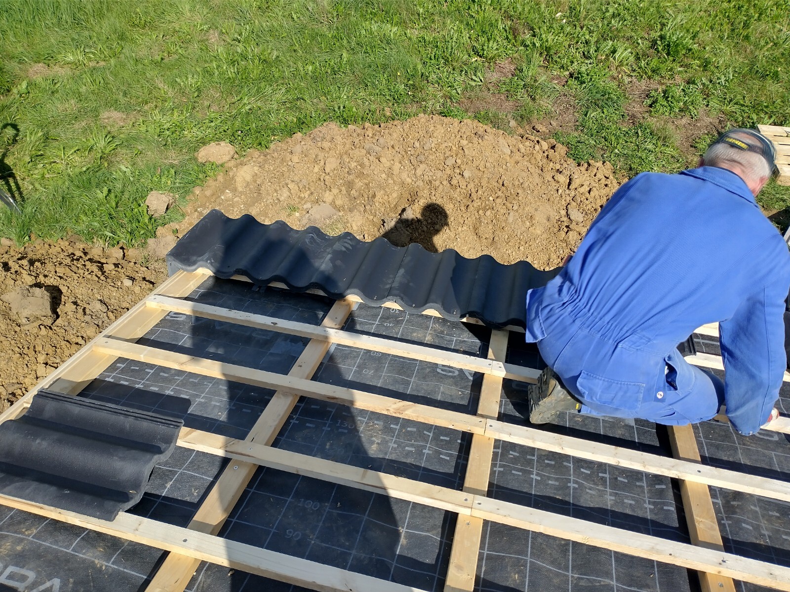 Bauarbeiter in blauem Overall montiert schwarze Wellblechplatten auf Holzunterkonstruktion.