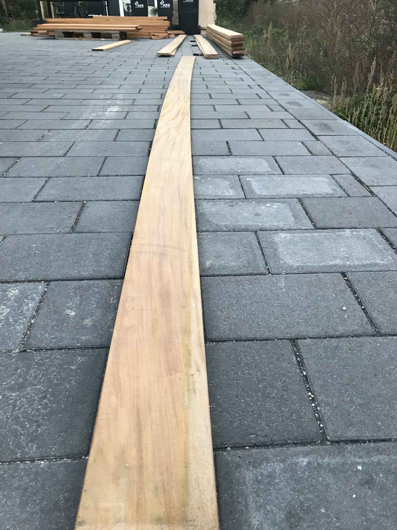 Lange Holzlatte liegt mittig auf grauen Pflastersteinen; Bauholzstapel im Hintergrund.