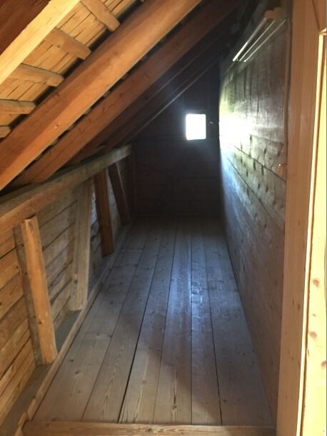 Dachboden mit Holzbalken, schrägen Decken und Holzdielen, kleines Fenster am Ende.