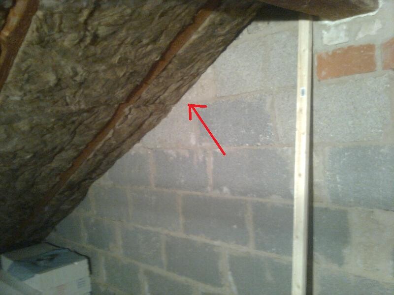 Dachboden mit schrägem Dach, Steinwand und Holzlatte; roter Pfeil markiert eine Ritze.