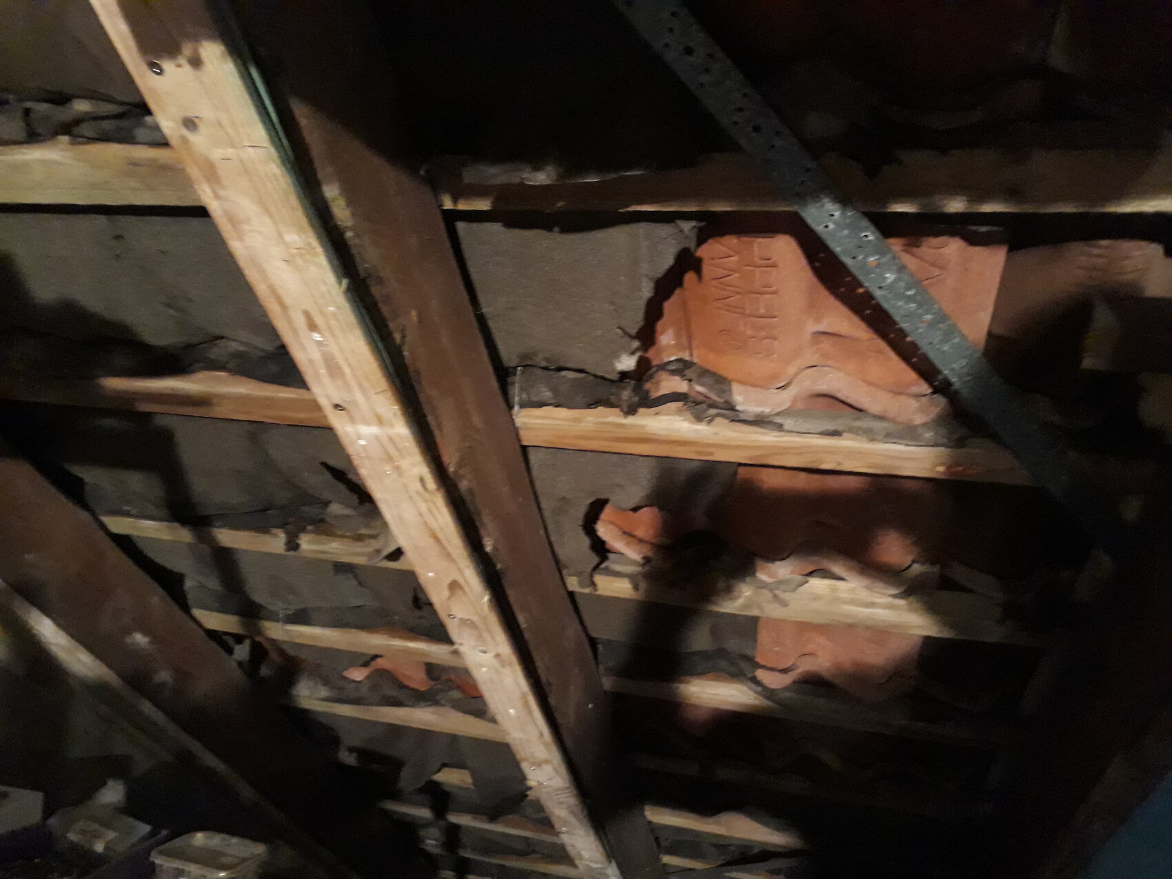 Dachboden: Holzbalken, rote Dachziegel, schwarze Dämmung und Metallband im Dunkeln