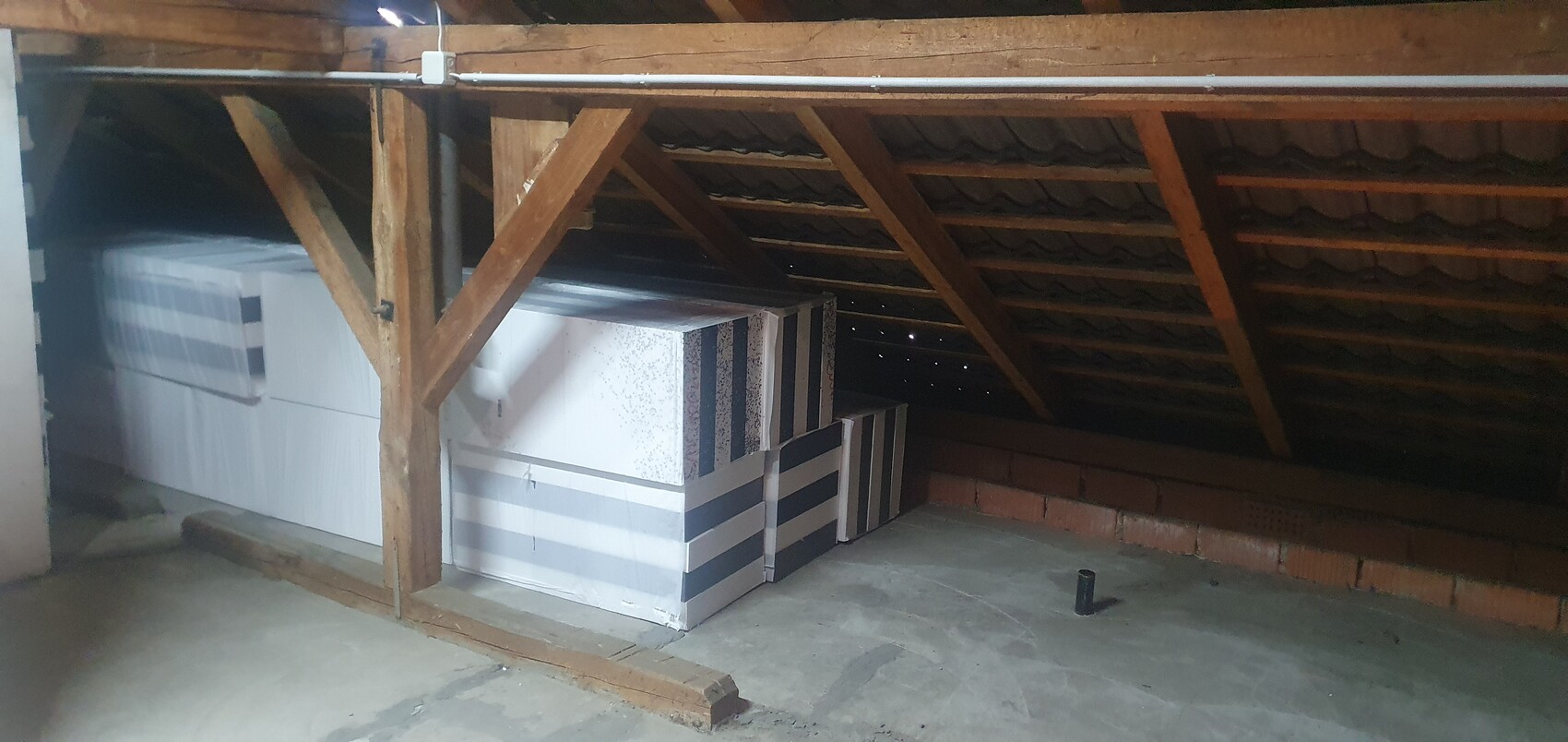 Dachboden mit Holzbalken; gestapelte weiße Isolierplatten mit grauen Streifen; Betonboden.
