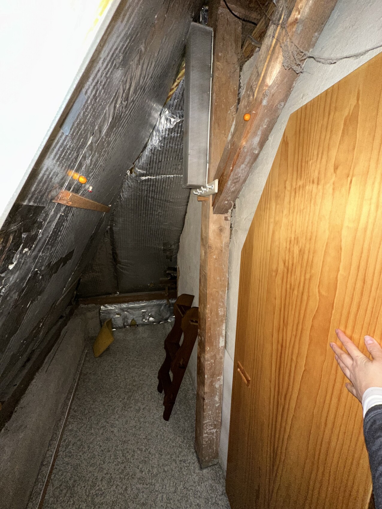 Enge Dachboden-Nische mit schrägem Dach, Holzbalken, Dämmung, Metallkanal, Tür rechts.
