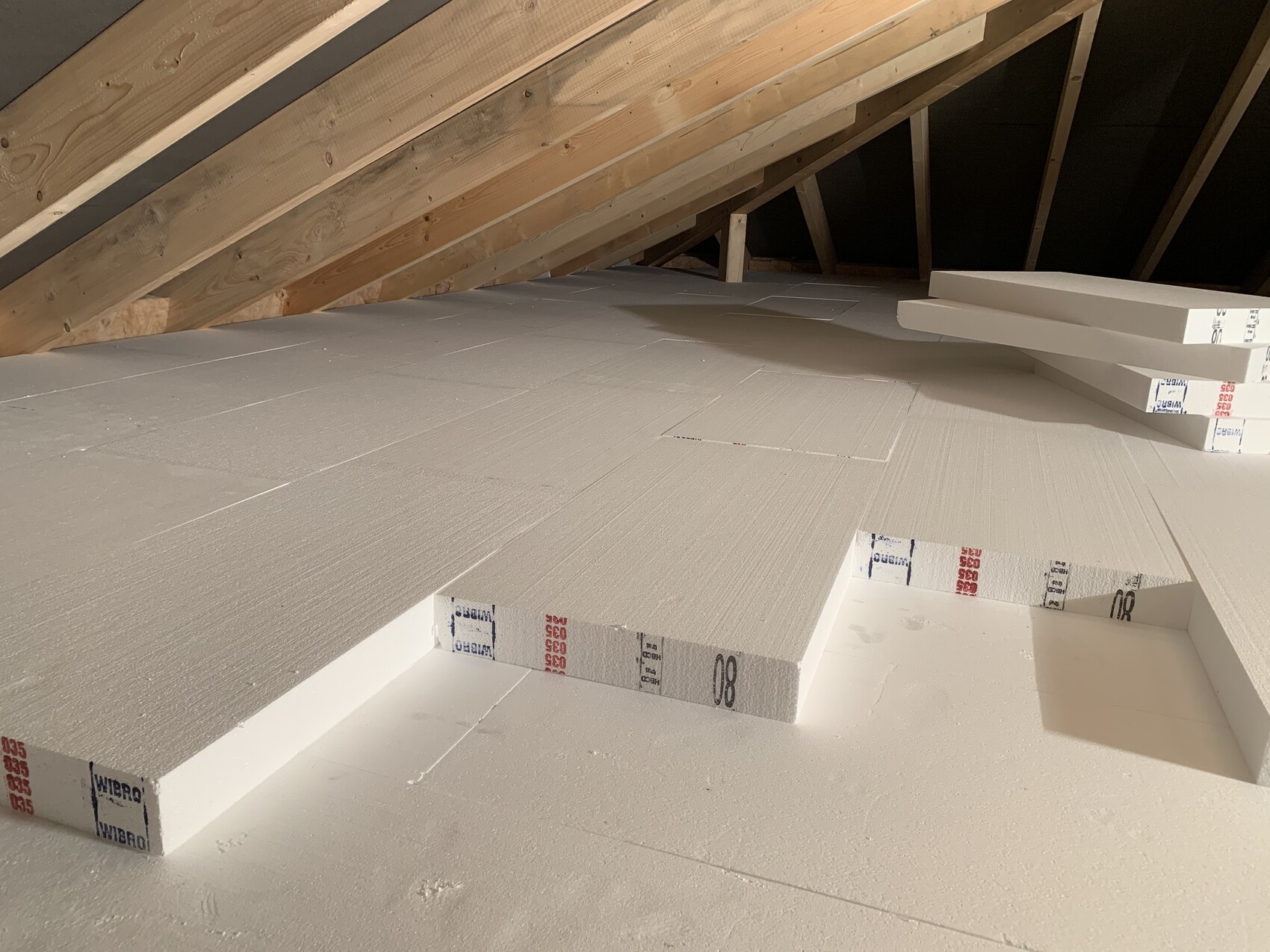 Dachboden mit weißen Isolierplatten auf dem Boden, Holz-Dachstuhl und Stapel Dämmplatten.