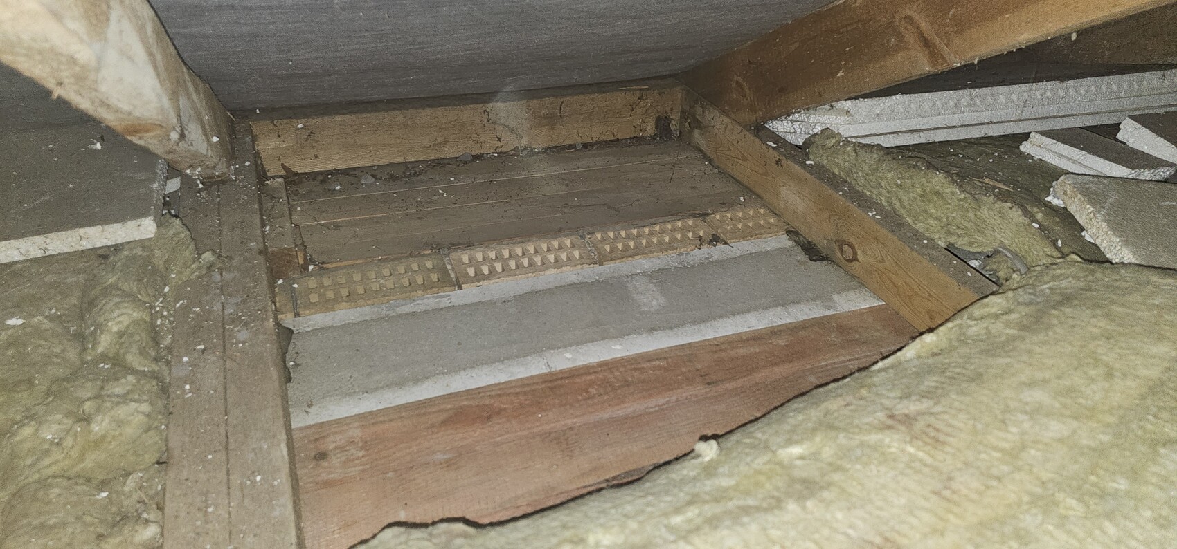 Dachboden mit Holzbalken, Dämmung und einem Bodenbelüftungsgitter.