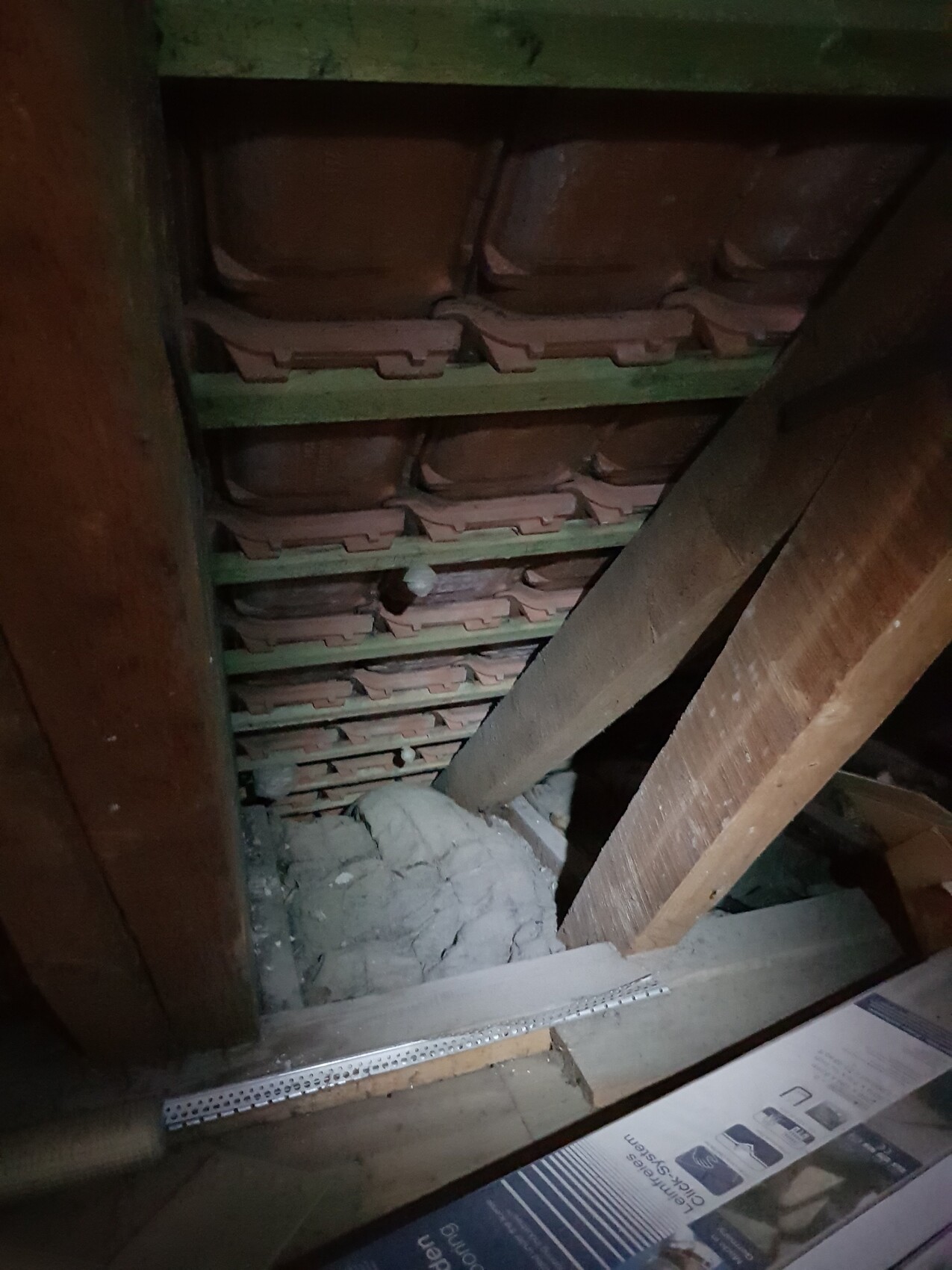 Dachboden mit Holzbalken, grünen Sparren, rotem Ziegelstapel und Dämmstoffresten.