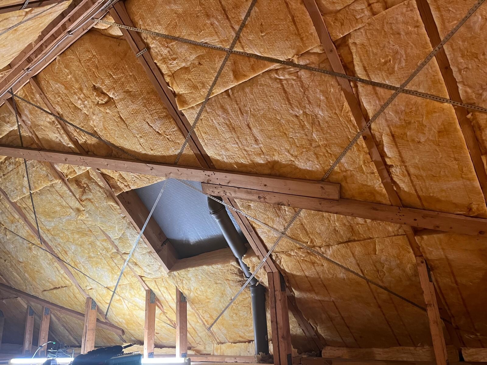 Dachboden mit gelbem Dämmstoff zwischen Holzbalken, Streben und grauem Rohr.