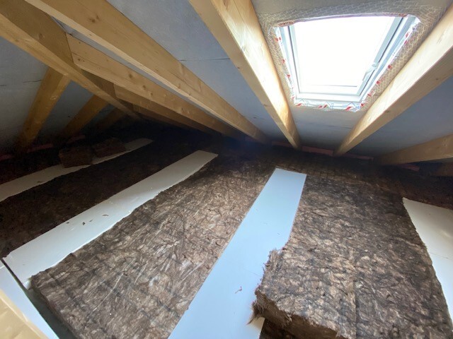 Dachboden mit Holzbalken, Dämmmaterial auf dem Boden und einem Dachfenster.
