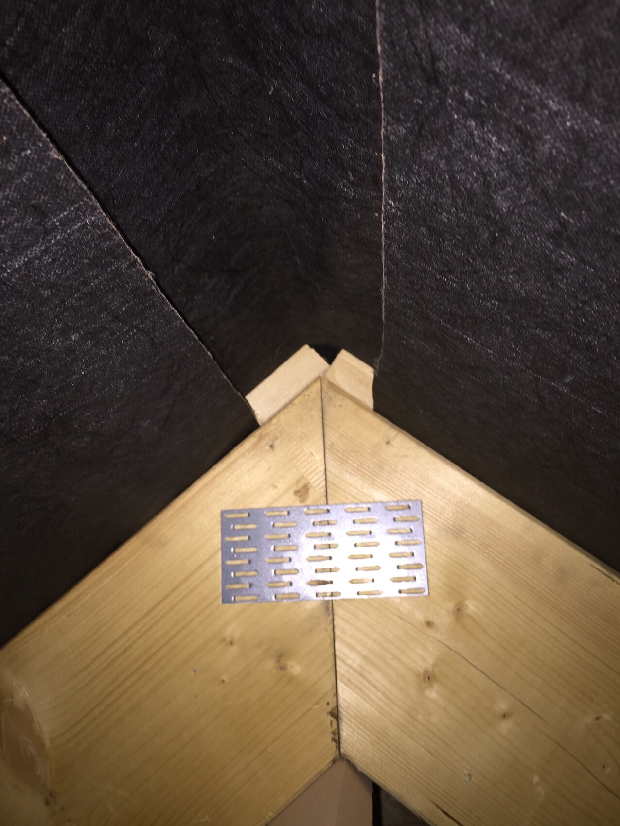 Dachboden-Ecke mit dunklen Wänden und hellem Holz; Metallgitter-Lüfter auf der Holzfläche.