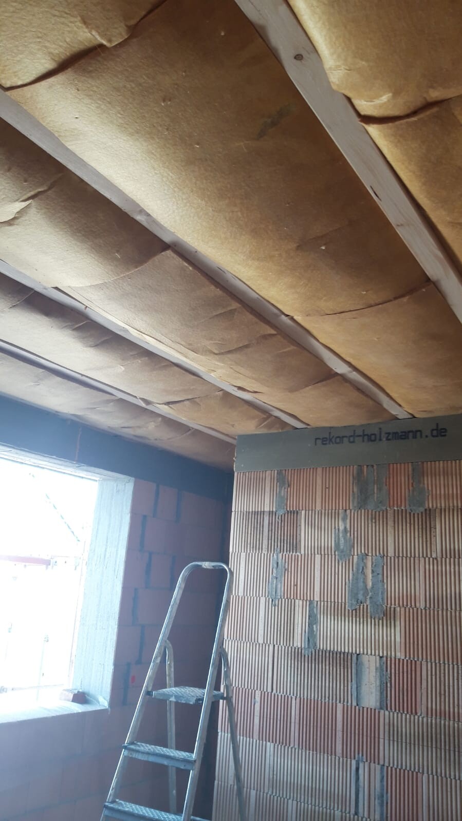 Dachboden-Decken-Dämmung zwischen Balken; Leiter und rohe Ziegelwand sichtbar.