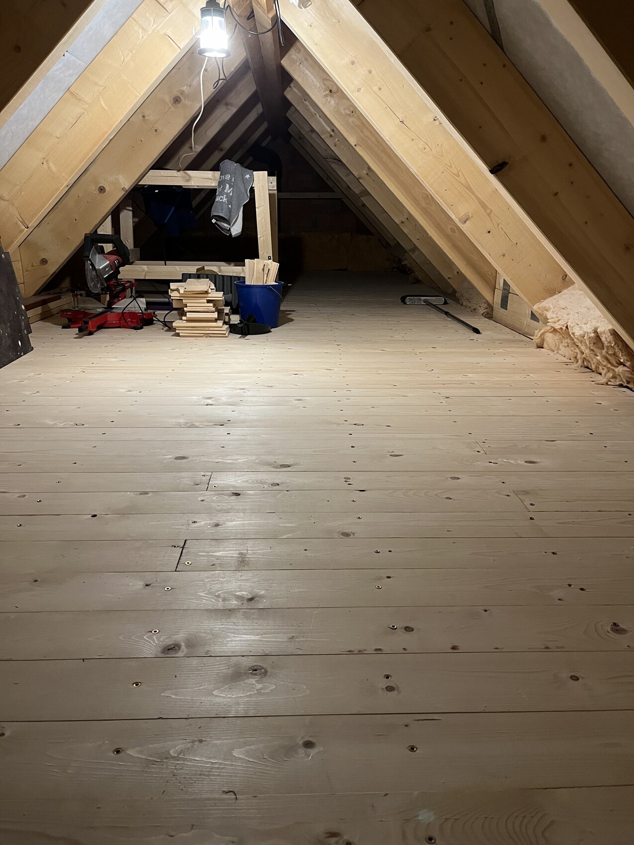 Dachboden mit Holzbalken, Bauholz, Werkzeug, Eimer, Holzplanken und Lampe.