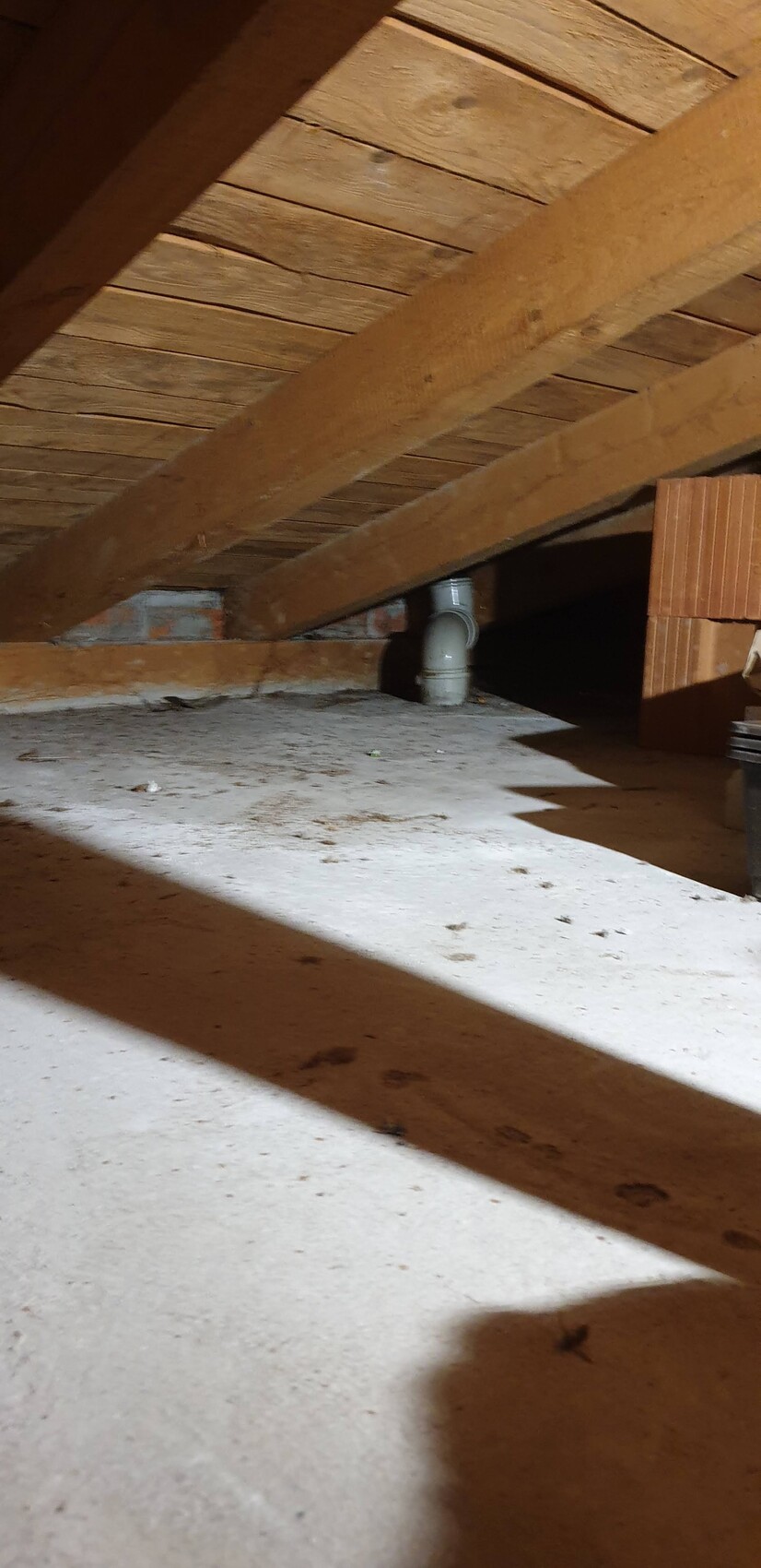 Dachboden mit Holzbalken, staubigem Boden, grauer PVC-Rohrverbindung und Ziegeln.