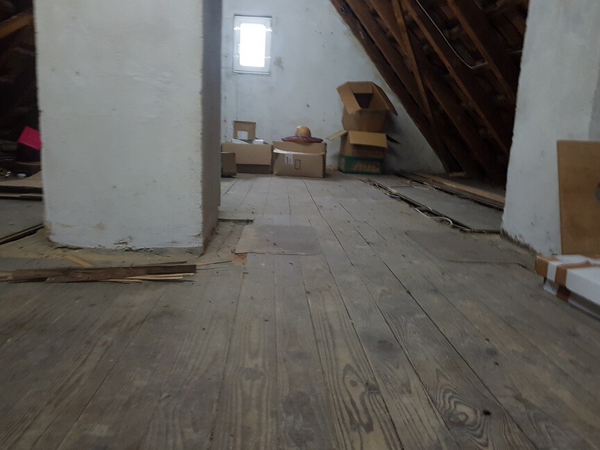 Dachboden mit Holzdielen, Kartons, Kisten und Bauholz; kleines Fenster im Hintergrund.
