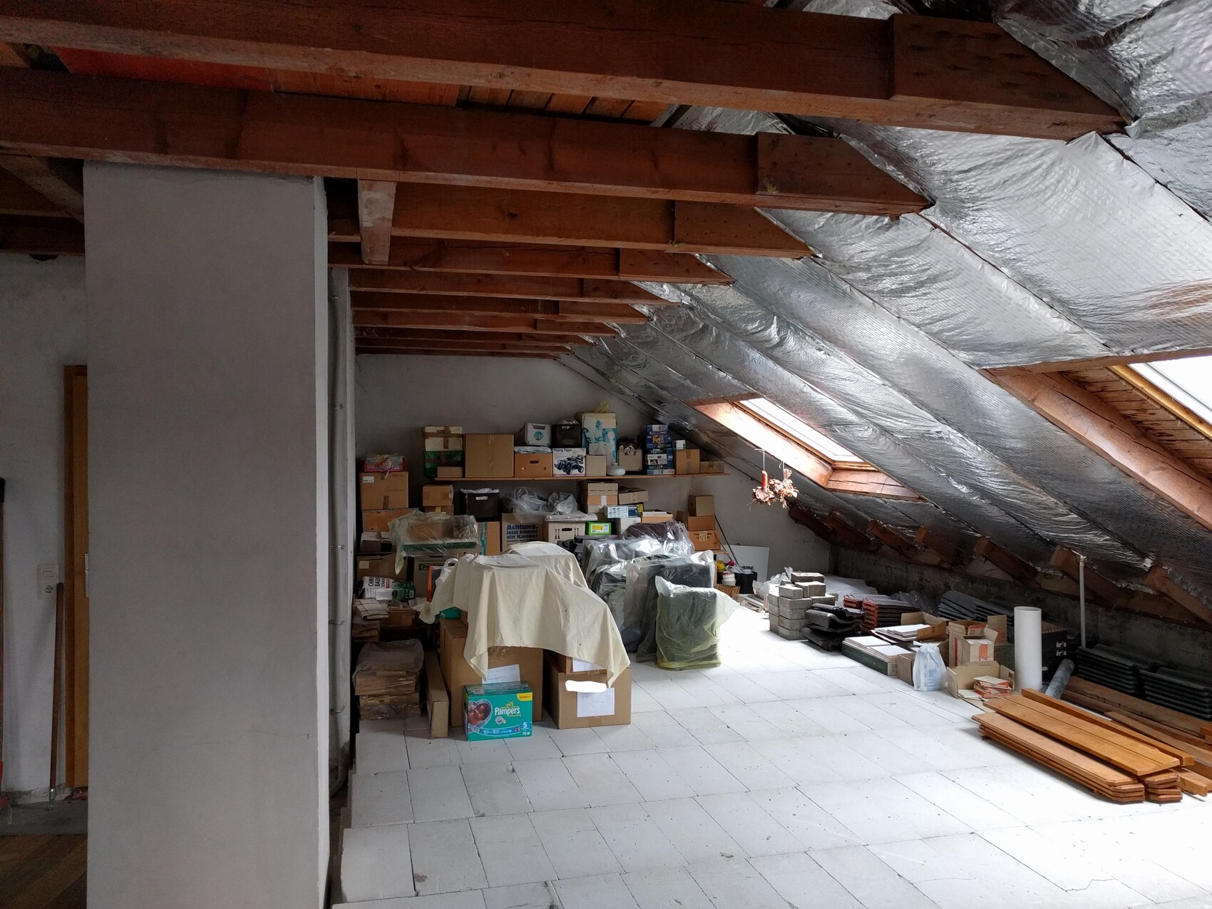 Dachboden mit Holzsparren, silberner Isolierfolie, viele Kartons, Kisten und Baumaterial stapelweise.
