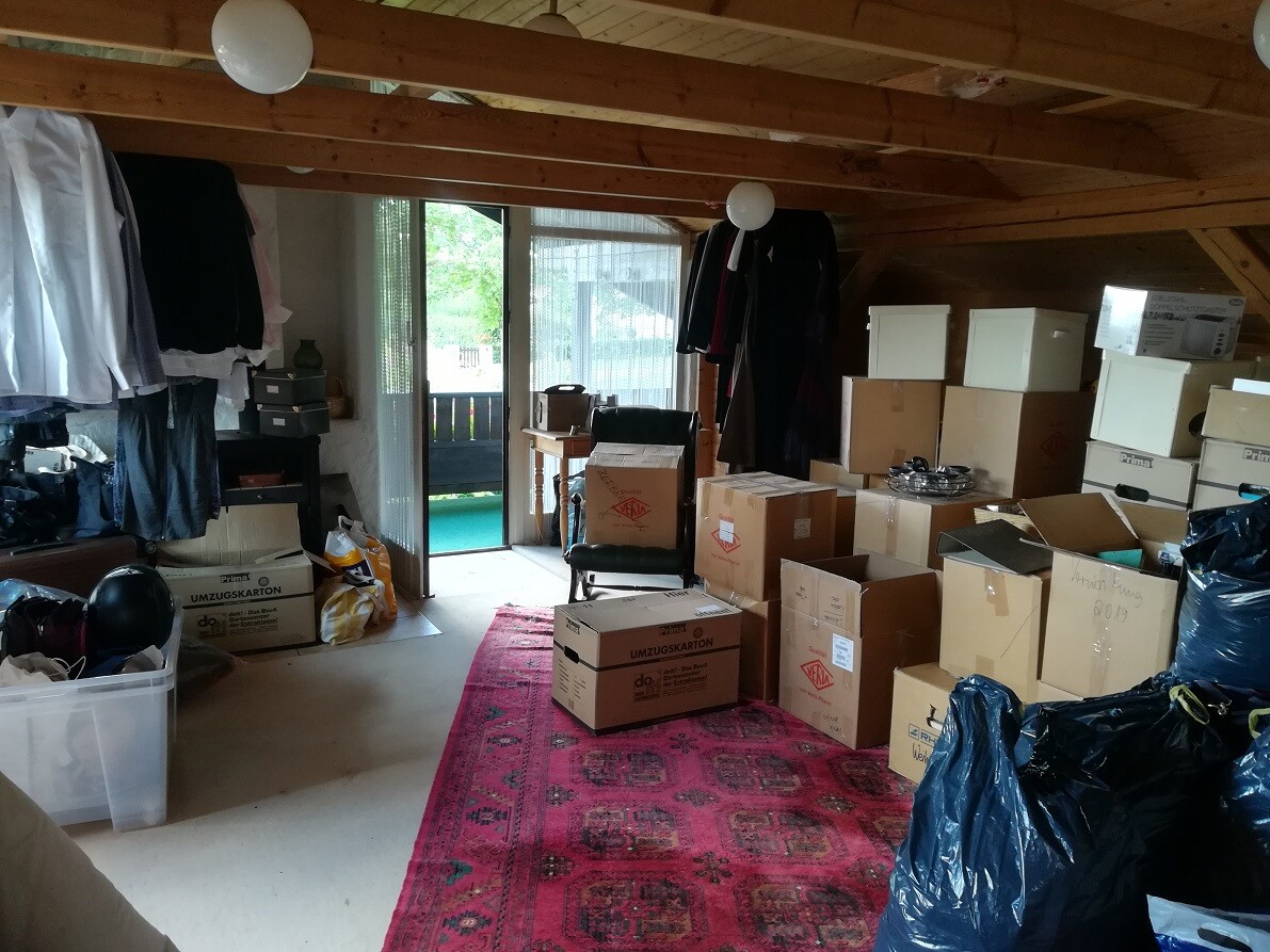 Dachboden-Abstellraum mit Kartons, Kleidung und offener Tür zum Garten.