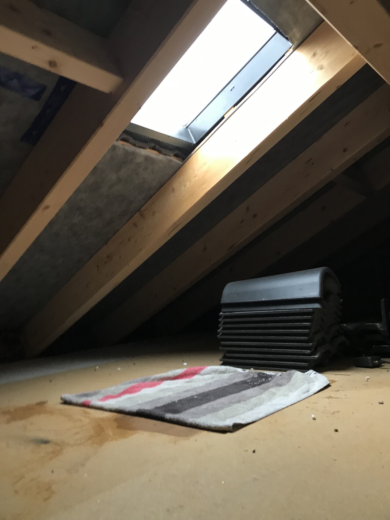Dachboden mit Holzbalken; Dachfenster lässt Licht herein; schwarze Lüftungsteile gestapelt.
