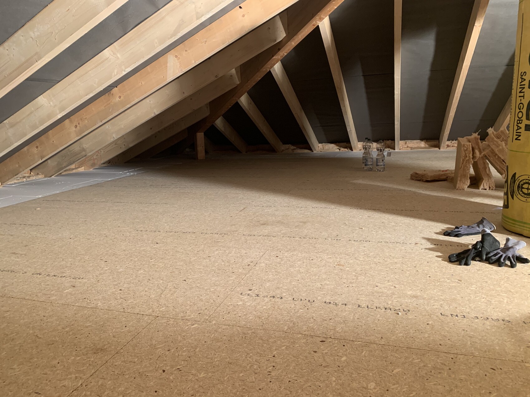 Dachboden im Rohbau: Dachstuhlbalken, Spanplattenboden, Handschuhe und Wasserflaschen.