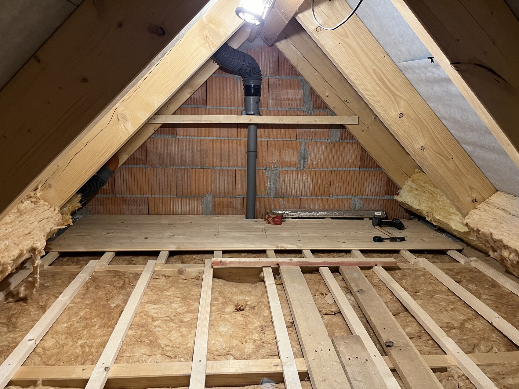 Dachboden mit Holzbalken, Backsteinwand, vertikalem Abflussrohr, Dämmung an Seiten, Werkzeuge.