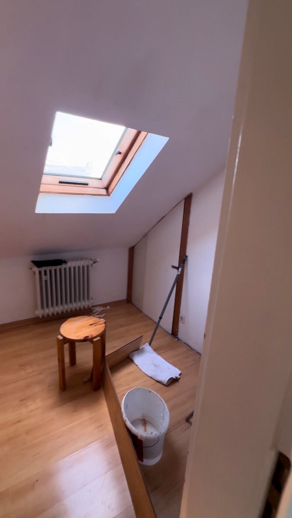 Kleines Dachbodenzimmer mit Dachfenster, Heizkörper, Hocker, Eimer und Mopp.