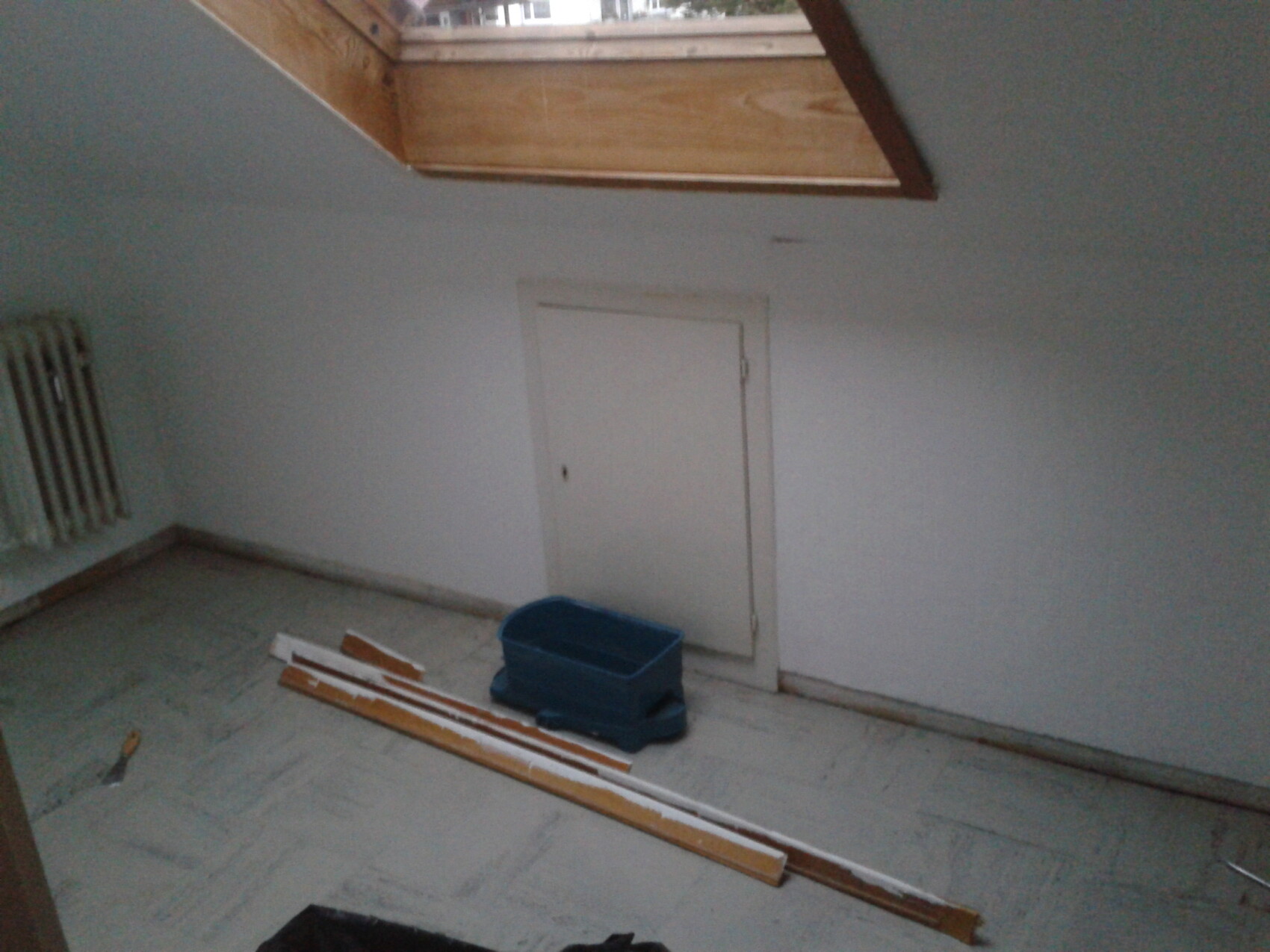 Kleiner Dachbodenraum mit Dachfenster, weißer Tür, Heizkörper links, Holzlatten und blauer Box.