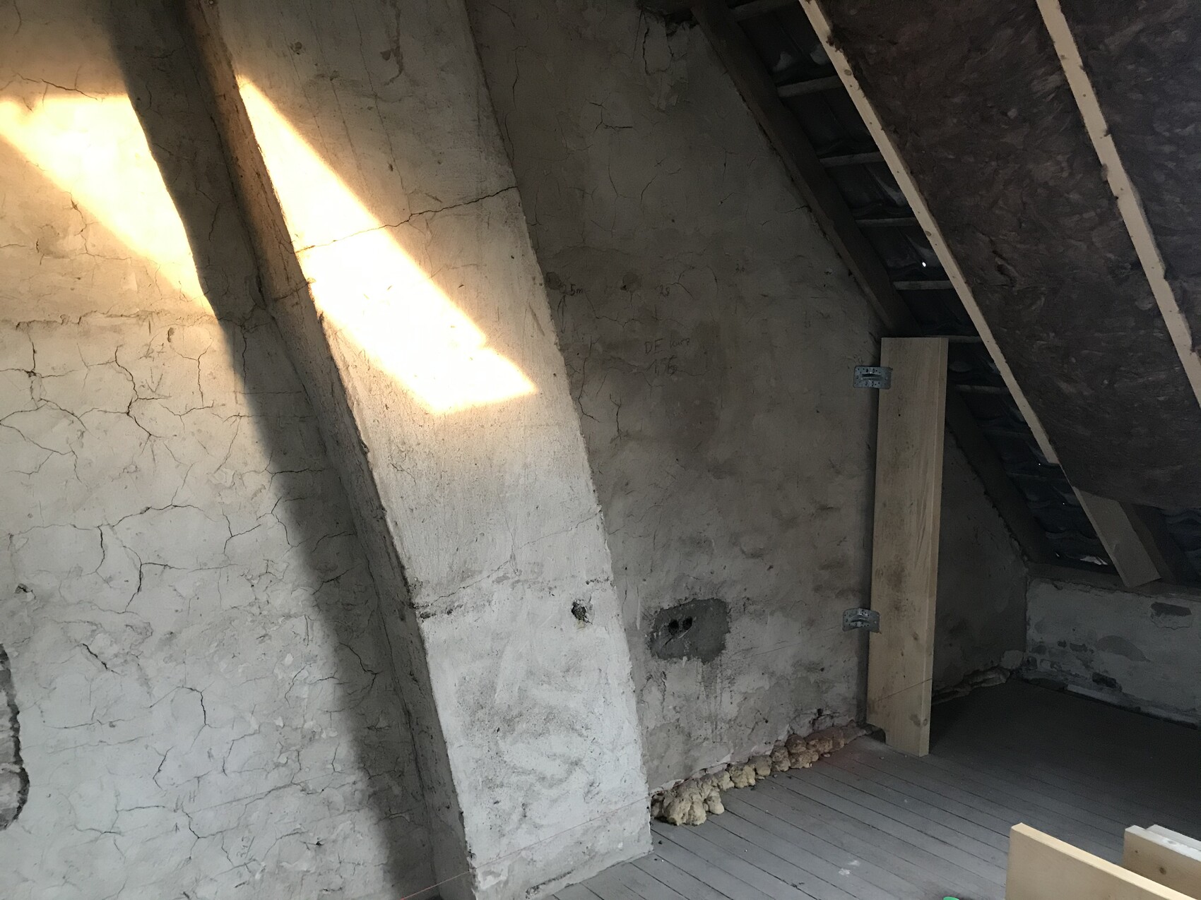 Dachboden mit rissiger Wand, diagonalem Stützpfeiler, Holzboden und Lichtstrahl durch Öffnung.