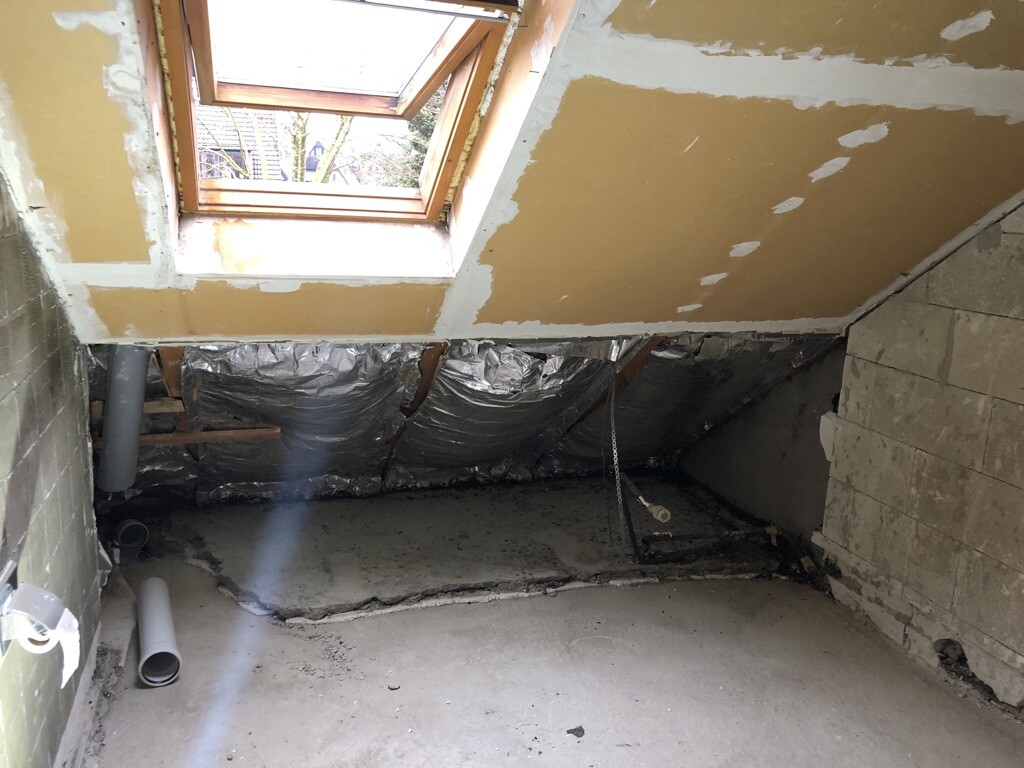 Dachgeschoss-Rohbau mit Dachfenster, Dämmfolie, Rohren und Betonboden.