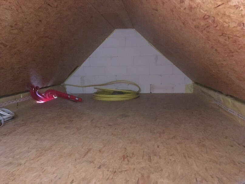 Dachboden mit schrägem Dach, weißer Backsteinwand im Hintergrund, gelber Schlauch und rotes Werkzeug links.