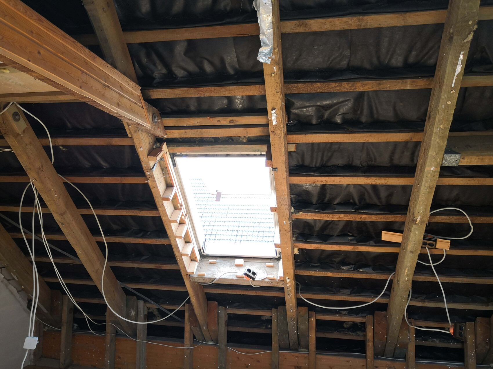 Dachboden mit offenem Lichtausschnitt, Holzbalken, schwarze Dämmung, Kabel.