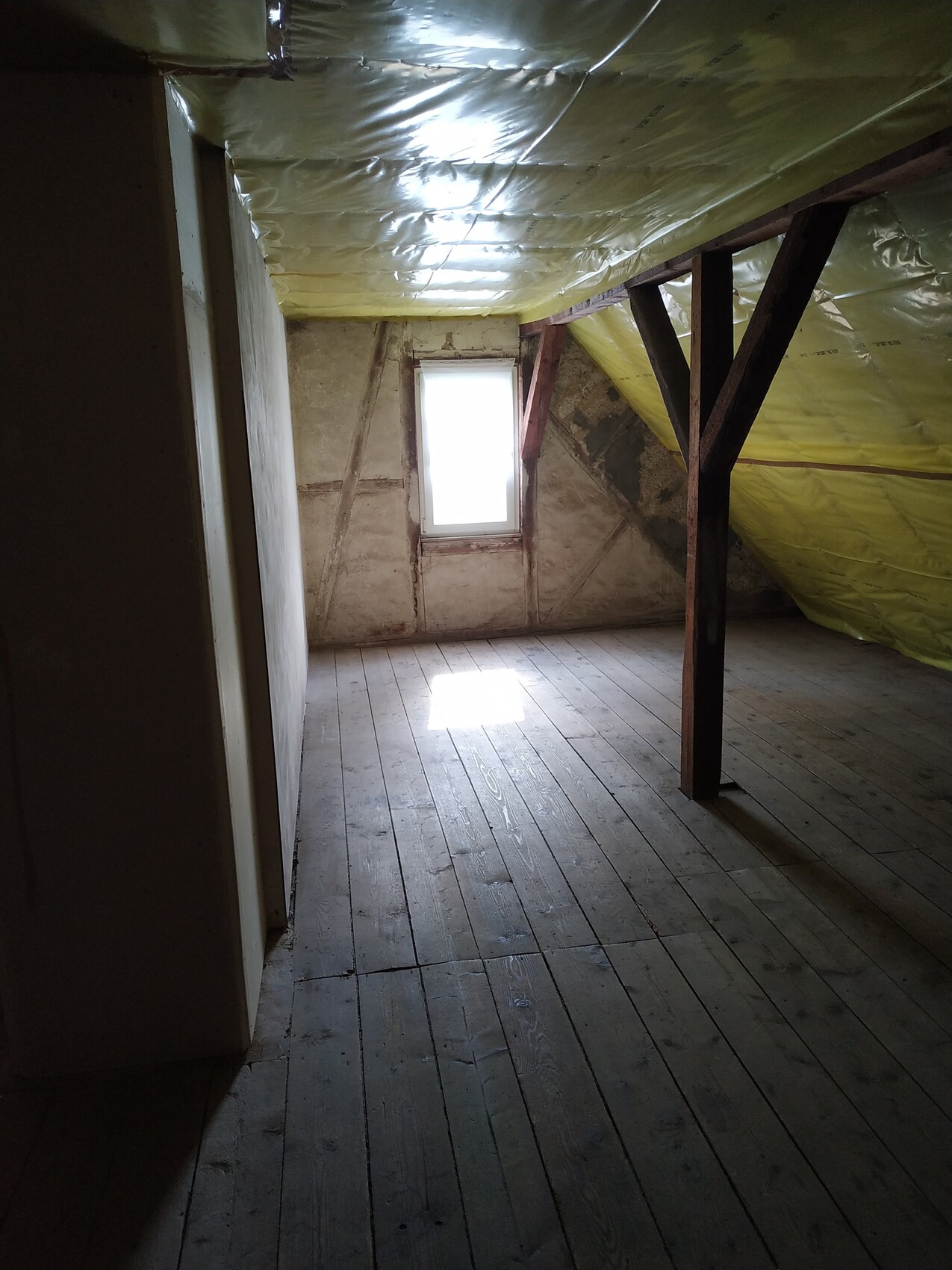 Dachbodenraum mit Holzdielen, dunklen Balken, gelber Dämmfolie an der Decke und kleinem Fenster.