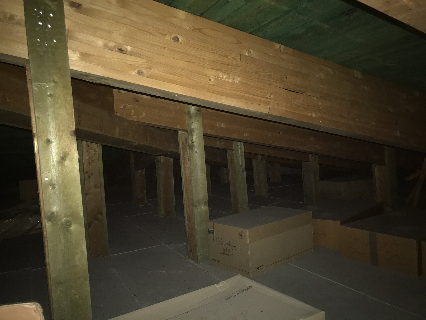 Dachboden mit Holzbalken, Stützpfosten und Kartons auf dem staubigen Boden.