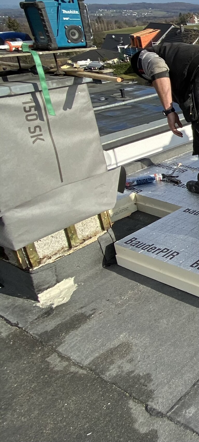 Arbeiter auf Flachdach installiert Dämmplatten; Werkzeug und BauderPIR-Material sichtbar.