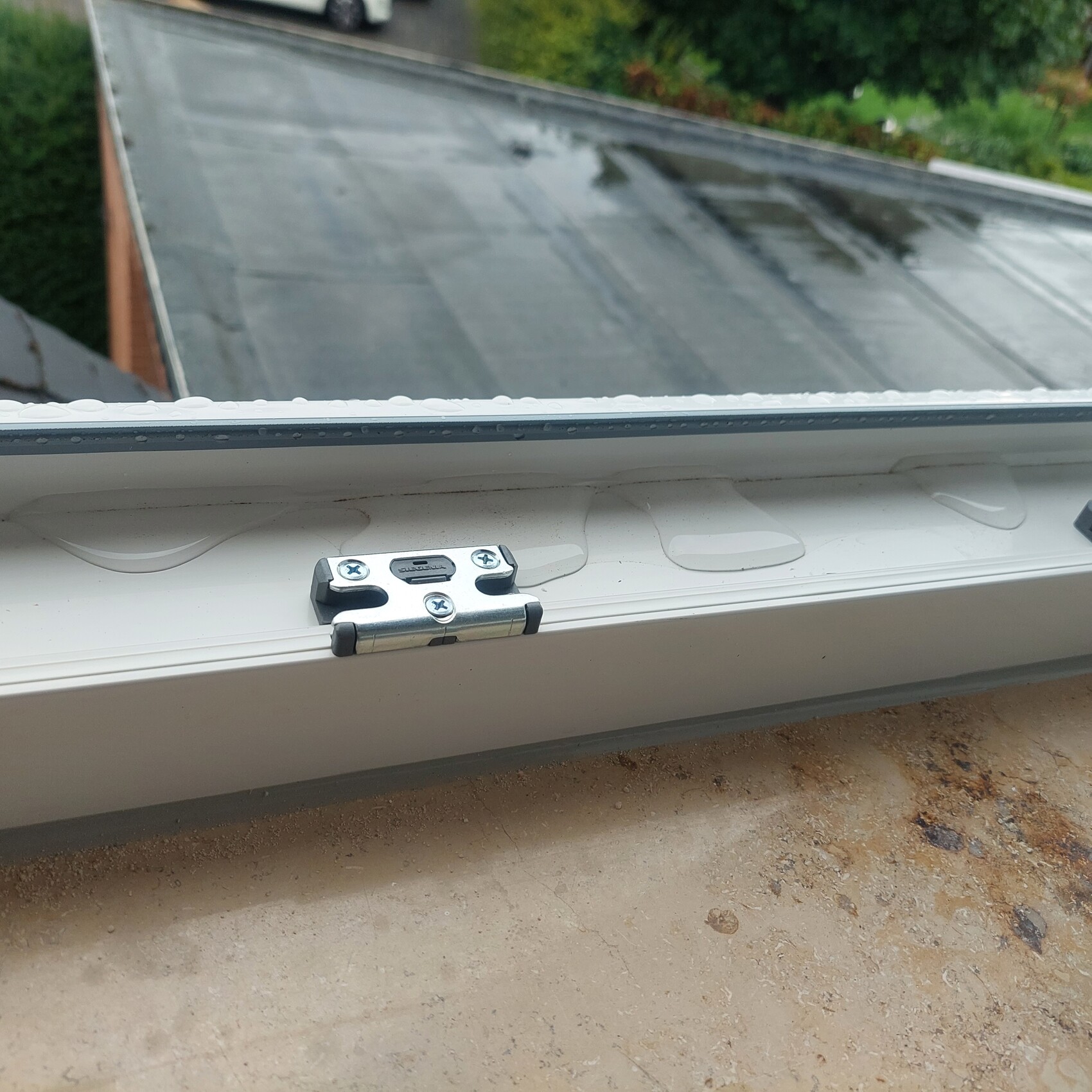 Dachfenster mit silbernem Metall-Scharnier auf weißem Rahmen, regennasses Glas.