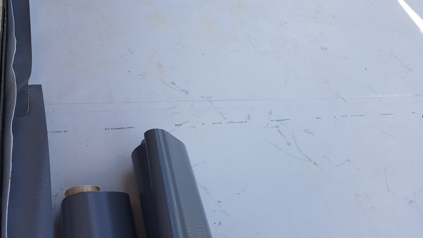Mehrere dunkle Rollen Dachbahnmaterialien liegen auf einer hellgrauen Unterlage, links.