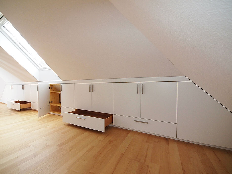 Weiße Einbauschränke unter schräger Dachdecke im Dachgeschoss, mit offenen Schubladen und Holzfußboden.