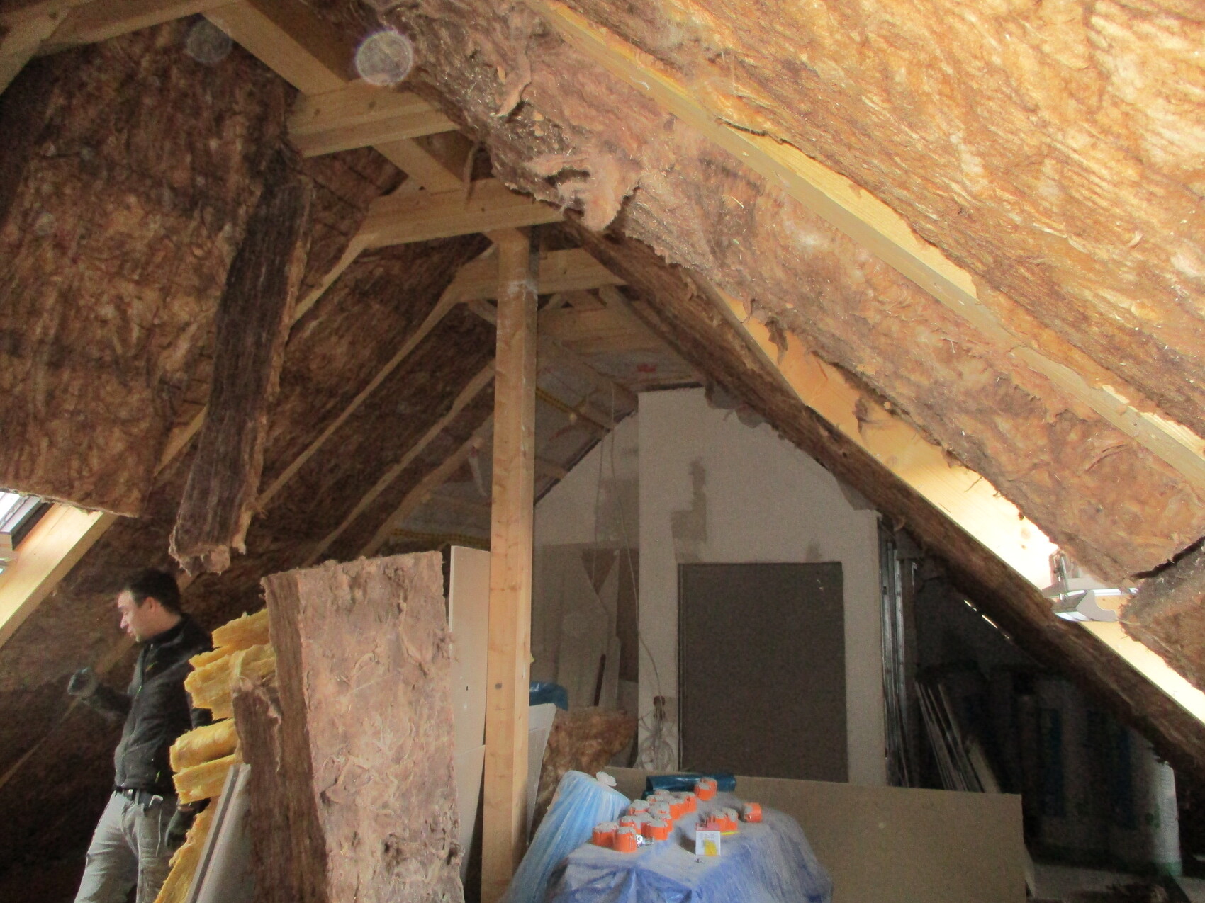Dachboden mit Holz-Dachstuhl und Dämmung, links stehender Bauarbeiter, Baumaterialien.